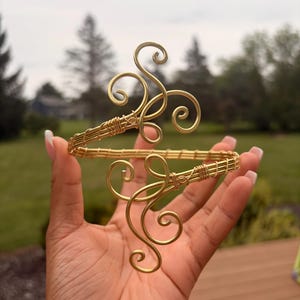 Puede incluir: Un brazalete de alambre de color dorado con un diseño de banda continua y dos motivos de volutas simétricos y prominentes en los extremos abiertos. El alambre de metal está enrollado firmemente alrededor de la estructura principal.
