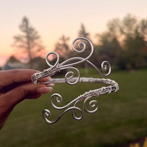 May include: A silver-colored wire cuff bracelet featuring a multi-strand band and two decorative ends shaped into intricate, swirling scroll patterns. The metal wire is tightly coiled at the connection points to secure the structure of the piece.