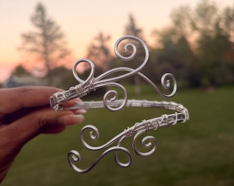 May include: A silver-colored wire cuff bracelet featuring a multi-strand band and two decorative ends shaped into intricate, swirling scroll patterns. The metal wire is tightly coiled at the connection points to secure the structure of the piece.