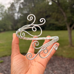 May include: A silver-colored wire cuff bracelet featuring a flexible, open-ended design. The two ends of the bracelet are shaped into intricate, swirling, scroll-like patterns, while the main band consists of multiple parallel wire strands.
