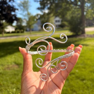 May include: A silver-colored wire arm cuff featuring an open design with intricate, swirling scrollwork patterns at both ends. The metal wire is tightly coiled and wrapped to form the structure, creating a continuous, flexible band for the arm.