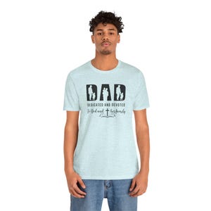 以下が含まれることがあります： ライトブルーの霜降りTシャツ。黒い文字で「DAD」と書かれ、その文字の中に親子のシルエットが描かれています。その下には「DEDICATED AND DEVOTED To God and his family」という文字と、開いた本の上に十字架のアイコンがデザインされています。