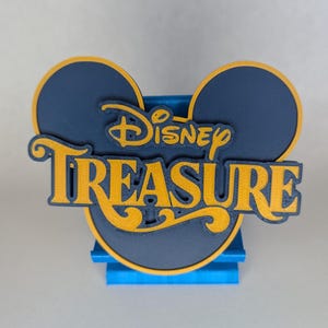May include: A navy blue and gold Disney Treasure logo in the shape of Mickey Mouse ears. The word "Disney" is in a script font above the word "Treasure". The logo is on a blue stand.