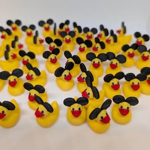 May include: A large collection of small yellow rubber ducks, each featuring a pair of black circular ears attached to the top of the head and a bright red beak. The ducks are arranged in a scattered pattern across a plain white surface.