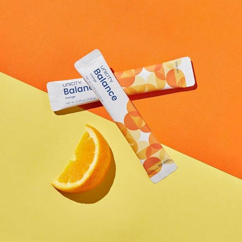 K&ouml;nnte beinhalten: Zwei wei&szlig;e Unicity Balance Orange-P&auml;ckchen mit orangefarbenen und gelben geometrischen Designs liegen auf einem gelben und orangefarbenen Hintergrund. Eine Orangenscheibe befindet sich in der linken unteren Ecke.