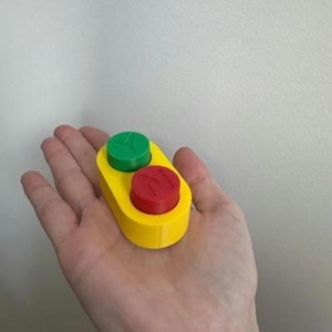 Puede incluir: Un pequeño objeto amarillo de forma ovalada con dos botones circulares en la parte superior. El botón superior es verde con la letra Y en relieve, y el botón inferior es rojo con la letra N en relieve. El objeto tiene un acabado mate liso.
