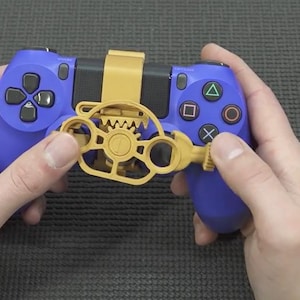 May include: A blue video game controller featuring a yellow plastic steering wheel attachment mounted over the center. The attachment includes a gear mechanism and a clip that secures it to the controller, positioned over the analog sticks.