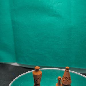 May include: Three small, brown, 3D-printed bottle-shaped objects with flat tops. Two of the objects are marked with the numbers 63 and 834, while the third is unmarked. These items are positioned on a reflective surface, creating a mirrored effect.