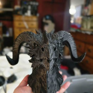 May include: A matte black 3D printed dragon bust featuring a detailed scaly texture, a sharp beak-like snout, and two large, curved ram-like horns extending from the sides of the head. The creature has a pointed crest and defined facial features.
