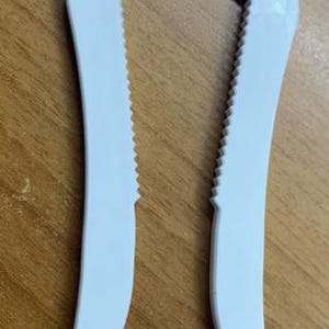 May include: A white plastic jar opener with a U-shaped design. The top section has a serrated edge, and the two arms have smooth, curved edges. The opener is designed to grip and twist off jar lids.