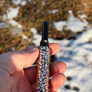 May include: A black and silver vaporizer pen encrusted with rhinestones. The pen has a white tip and is held in a hand against a blurred background of snow and brown grass.