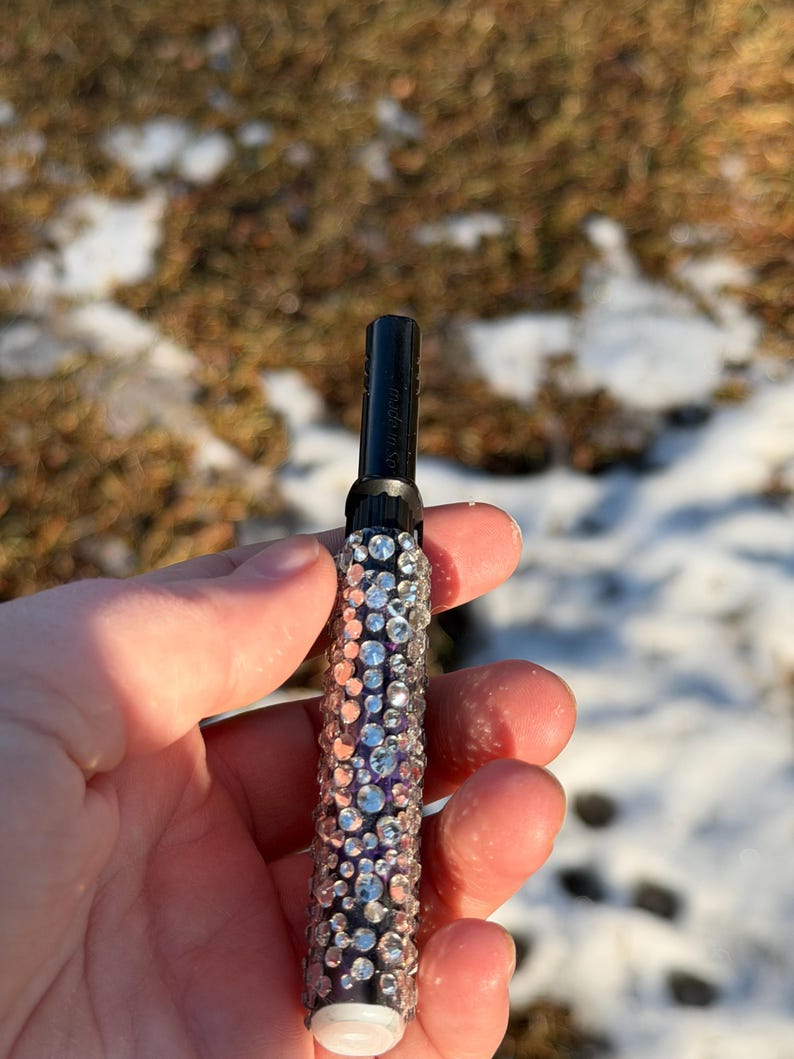May include: A black and silver vaporizer pen encrusted with rhinestones. The pen has a white tip and is held in a hand against a blurred background of snow and brown grass.