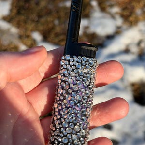 May include: A black lighter bedazzled with clear rhinestones of varying sizes. The rhinestones are arranged on a purple background. The lighter has a black top and a white base. The lighter is held in a hand.
