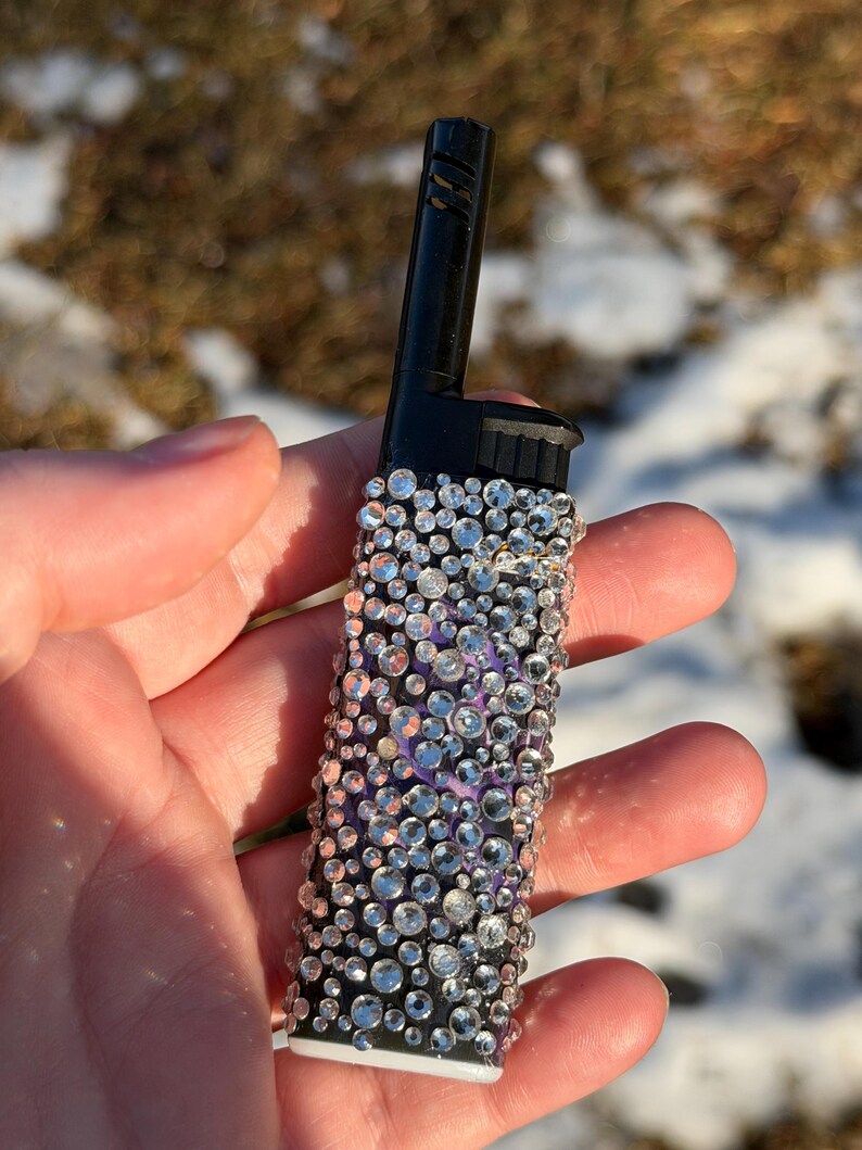 May include: A black lighter bedazzled with clear rhinestones of varying sizes. The rhinestones are arranged on a purple background. The lighter has a black top and a white base. The lighter is held in a hand.