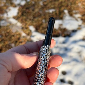 May include: A black lighter with a bedazzled design. The lighter is covered in a pattern of clear and silver rhinestones. The background is a blurred view of a snowy outdoor scene.