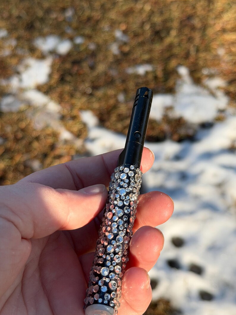 May include: A black lighter with a bedazzled design. The lighter is covered in a pattern of clear and silver rhinestones. The background is a blurred view of a snowy outdoor scene.