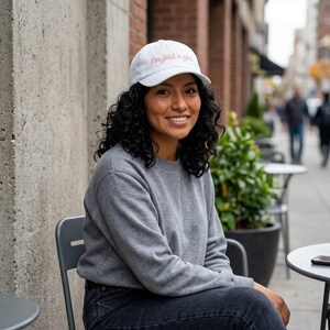 Peut inclure: Une casquette de baseball blanche avec le texte brodé "I'm just a girl" en écriture cursive rose, associée à un sweat-shirt gris à manches longues à col rond et un jean bleu foncé, formant une tenue décontractée et neutre.