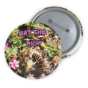May include: A round button with the text "EAT THE RICH" in pink letters. The button's design features a close-up of a caterpillar on a bed of green plants, dirt, and small pink flowers. The button has a silver pin back.