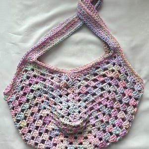 Puede incluir: Bolso de crochet hecho a mano en colores pastel, incluyendo rosa, morado, azul y amarillo. El bolso presenta un patrón de punto de la abuela y una correa larga para llevar al hombro. El bolso es de diseño suave y flexible.