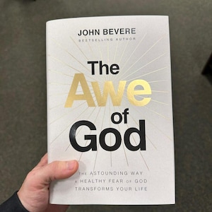 May include: A hardcover book titled "The Awe of God" by John Bevere. The cover is white with gold lettering and radiating lines. The text "The Awe of God" is prominently displayed. The book's subtitle reads, "The Astounding Way a Healthy Fear of God Transforms Your Life."