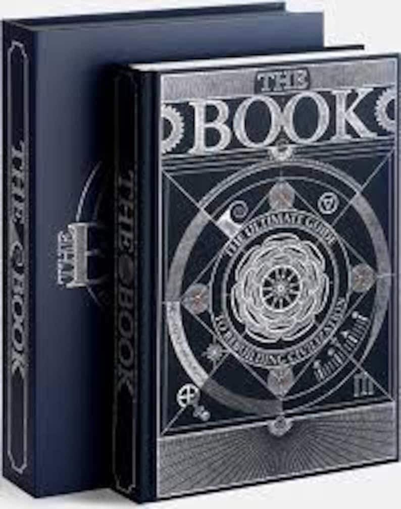 Op de afbeelding: Twee hardcover boeken met donkerblauwe covers. Het boek rechts heeft de titel "THE BOOK" in zilveren letters, met ingewikkelde geometrische ontwerpen. De rug van het boek links leest ook "THE BOOK".