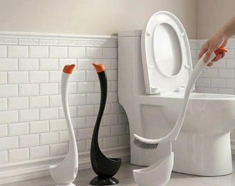 May include: Swan-shaped toilet brush and holder set. The set includes a white brush with an orange accent, a black holder, and a white holder. The brush is being held near a white toilet.