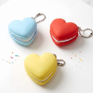 Può includere: Tre portachiavi a forma di cuore in blu, rosso e giallo, simili a macarons. Ogni portachiavi ha una catena argentata e un ripieno bianco tra le due metà del cuore. Ci sono delle decorazioni sparse.