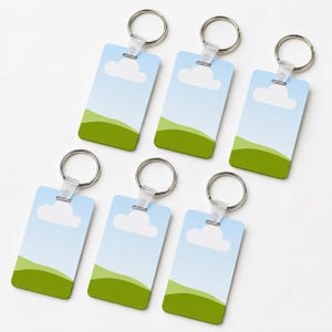 Puede incluir: Un conjunto de seis llaveros rectangulares con un diseño de paisaje impreso. Cada llavero muestra un cielo azul claro con una nube blanca sobre una colina verde, unido a una anilla de metal plateado mediante un conector de plástico transparente.