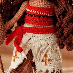 May include: A handcrafted crochet doll featuring long wavy brown yarn hair, wearing a bright red crop top with black zigzag embroidery and a white skirt with a red waist tie, orange cross-stitch details, and long white fringe along the bottom hem.