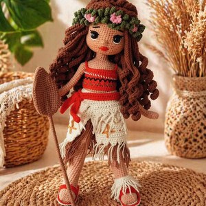 May include: A crocheted doll with long brown curly hair, wearing a green floral crown, a red patterned crop top, and a white fringed skirt with orange cross embroidery. The doll holds a brown crocheted paddle and wears red sandals with white fringe.