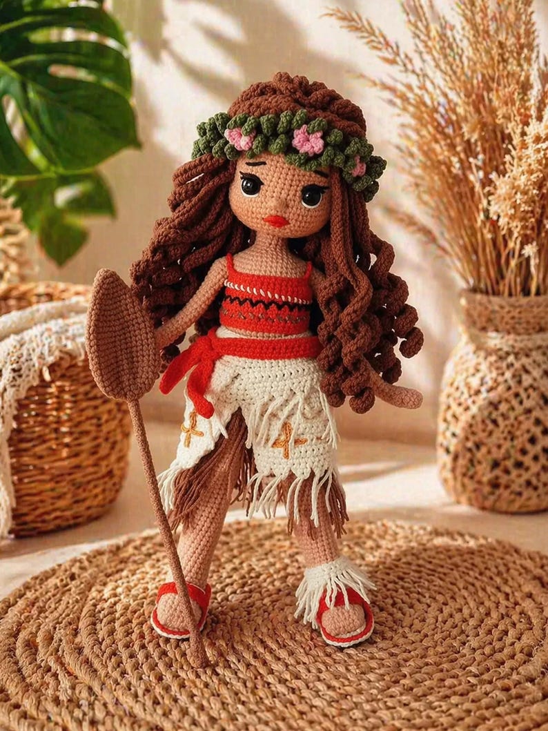 May include: A crocheted doll with long brown curly hair, wearing a green floral crown, a red patterned crop top, and a white fringed skirt with orange cross embroidery. The doll holds a brown crocheted paddle and wears red sandals with white fringe.