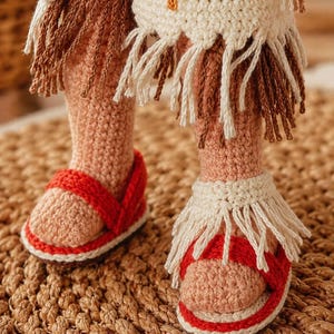 May include: A pair of crocheted doll legs wearing red sandals with white soles and cream-colored fringe accents. The doll is dressed in a cream-colored crocheted garment featuring brown and cream fringe along the hem and orange embroidery.