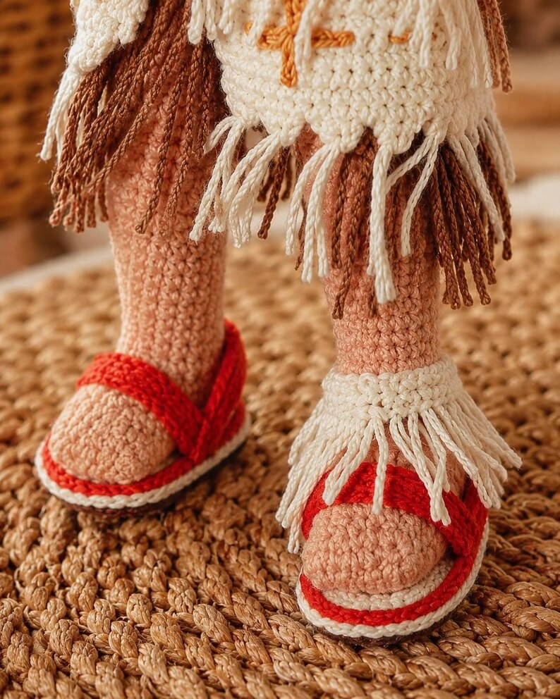 May include: A pair of crocheted doll legs wearing red sandals with white soles and cream-colored fringe accents. The doll is dressed in a cream-colored crocheted garment featuring brown and cream fringe along the hem and orange embroidery.