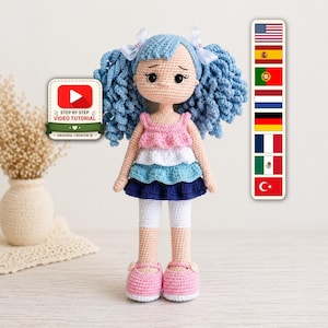 Op de afbeelding: Een handgemaakte gehaakte pop met blauw garen haar, gekleed in een roze, witte, blauwe en marineblauwe jurk, witte leggings en roze schoenen. De pop heeft een verdrietige uitdrukking. Een YouTube video tutorial logo is zichtbaar.