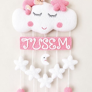 Puede incluir: Un colgante de pared de ganchillo con una nube blanca con cara dormida, mejillas rosadas y una pequeña muñeca con traje rosa encima. Una placa rectangular rosa muestra el nombre TUSEM en blanco, con estrellas blancas, una luna y esferas rosas colgando.