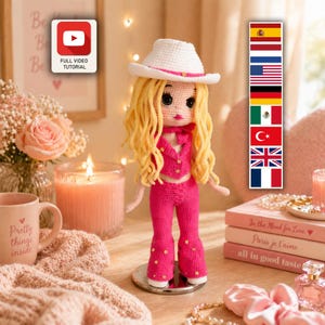 Puede incluir: Una muñeca de ganchillo con largo cabello rubio que lleva un sombrero de vaquero blanco, un chaleco rosa y pantalones acampanados a juego. La muñeca tiene pequeños detalles de flores amarillas en la ropa y se apoya en una base plateada.