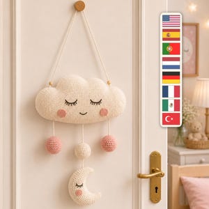 Op de afbeelding: Een crèmekleurige gehaakte wandhanger met een wolk met een slapend gezicht en roze wangen. Drie koorden hangen aan de wolk, elk eindigend in een kleine bol, met een halve maan met een slapend gezicht bevestigd aan het middelste koord.