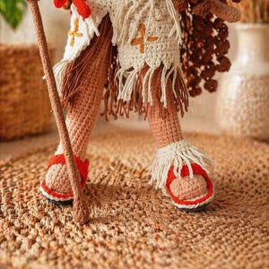 May include: A crocheted doll figure featuring tan skin, wearing a cream-colored skirt with fringe and orange cross-stitch details, a red waist tie, and red and white sandals with fringe. The figure holds a brown crocheted staff in one hand.