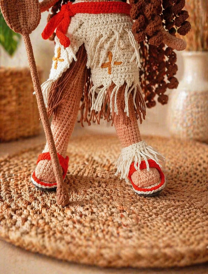 May include: A crocheted doll figure featuring tan skin, wearing a cream-colored skirt with fringe and orange cross-stitch details, a red waist tie, and red and white sandals with fringe. The figure holds a brown crocheted staff in one hand.