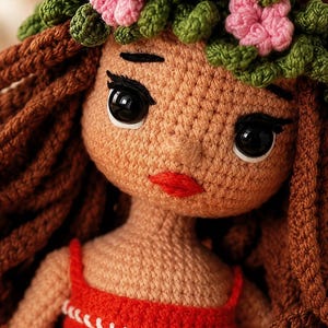May include: A handcrafted crochet doll featuring tan yarn skin, long brown yarn hair, and a green and pink floral crown. The doll has large black plastic eyes, embroidered black eyebrows, and red yarn lips. It wears a red top with white stitching.