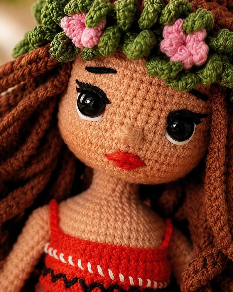 May include: A handcrafted crochet doll featuring tan yarn skin, long brown yarn hair, and a green and pink floral crown. The doll has large black plastic eyes, embroidered black eyebrows, and red yarn lips. It wears a red top with white stitching.