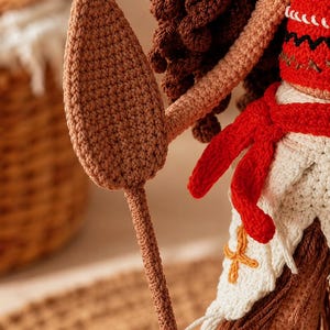 May include: A crocheted doll with dark brown curly hair, wearing a red top with black zigzag patterns and a white skirt with a red sash and orange cross embroidery. The doll holds a long, light brown crocheted spear with a pointed, leaf-shaped tip.