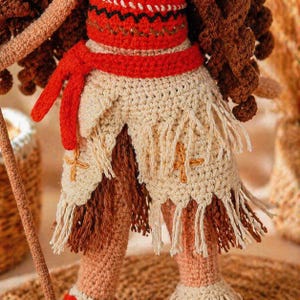 May include: A crocheted doll featuring a red top with black detailing, a red waist tie, and a cream-colored skirt with fringe and embroidered cross accents. The doll has dark brown curly hair and is holding a long, thin, light brown staff.