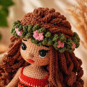 May include: A crocheted doll with long brown yarn hair and a floral crown made of green leaves and small pink flowers. The doll features large black eyes, a small nose, and bright red lips, wearing a red top with a white and black patterned trim.