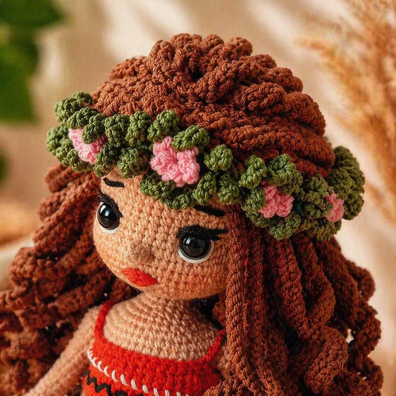 May include: A crocheted doll with long brown yarn hair and a floral crown made of green leaves and small pink flowers. The doll features large black eyes, a small nose, and bright red lips, wearing a red top with a white and black patterned trim.