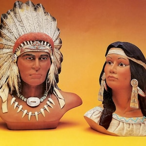 May include: Two ceramic busts of Native American figures. One figure wears a feathered headdress with red and white accents, a turquoise necklace, and a silver pendant. The other figure has long black hair, a headband, and feather earrings.
