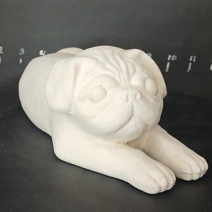 May include: White ceramic pug figurine, lying down with its head turned to the side. The figurine is unpainted and ready to be decorated.