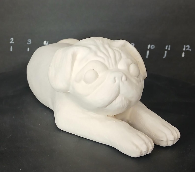 May include: White ceramic pug figurine, lying down with its head turned to the side. The figurine is unpainted and ready to be decorated.
