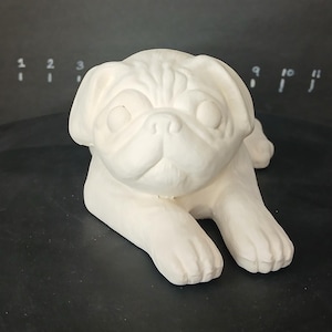 May include: White ceramic pug figurine, lying down with its head turned to the side. The figurine has a detailed face with a short snout and large eyes.