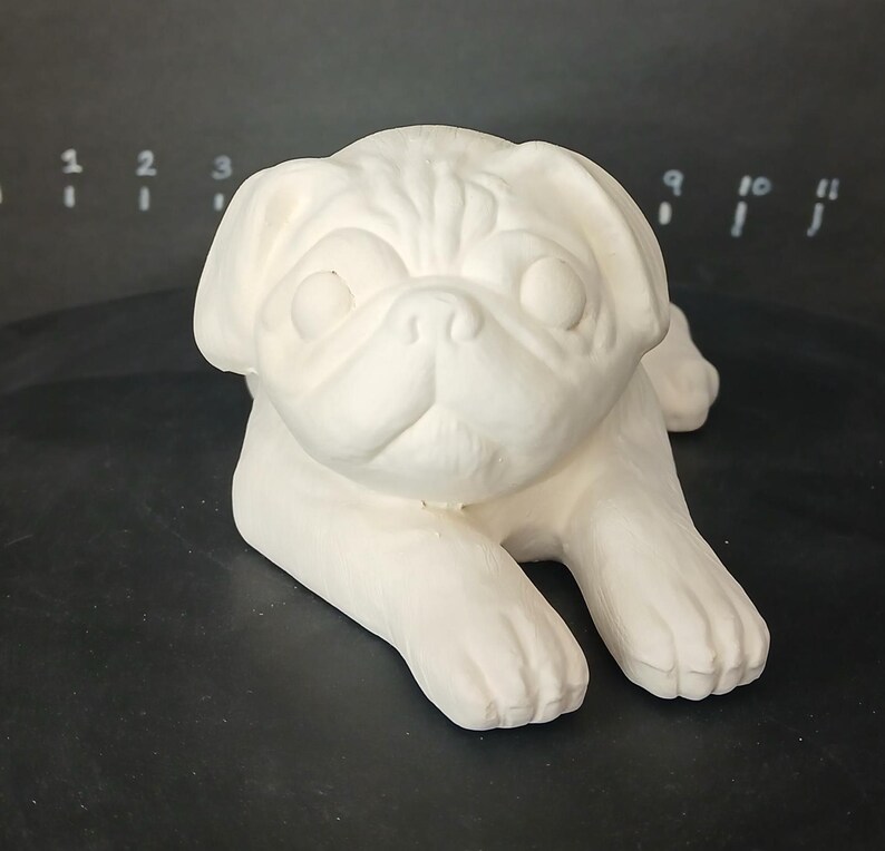 May include: White ceramic pug figurine, lying down with its head turned to the side. The figurine has a detailed face with a short snout and large eyes.